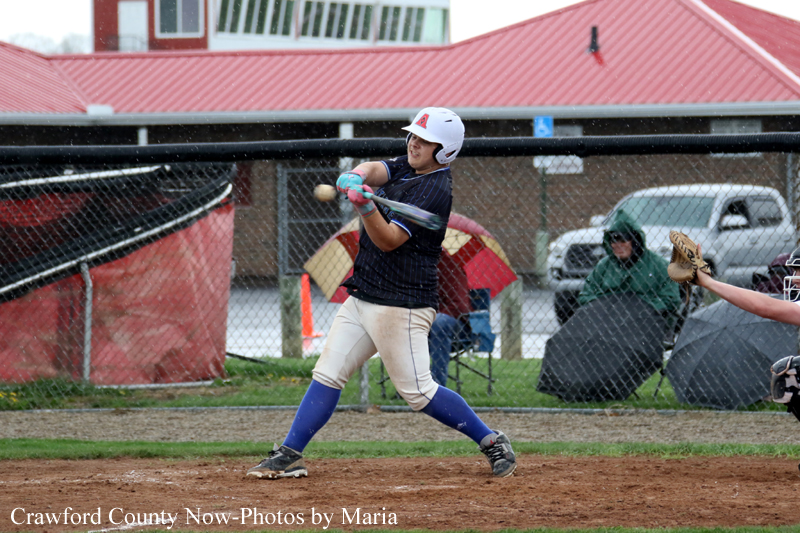 Baseball batter in a white helmet and navy jersey swings at a pitch on a dirt field, with a catcher and spectators behind a chain-link fence.