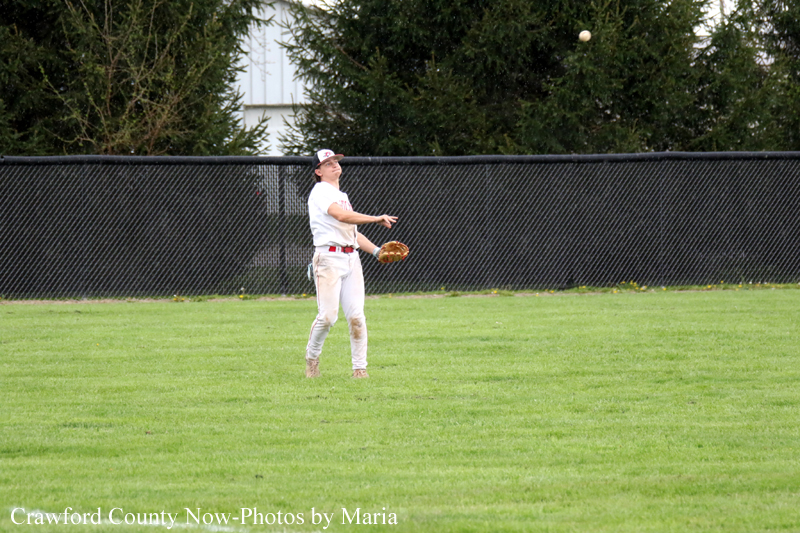 Baseball player in a white uniform on a grassy outfield, glove ready, ball descending toward him with a chain-link fence and trees behind.
