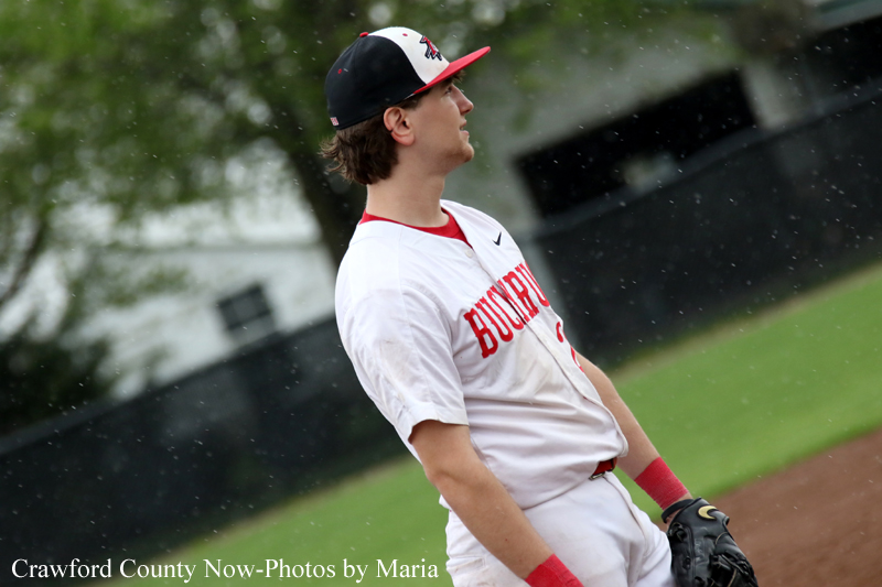 Baseball pitcher in a white and red uniform standing on a damp field, looking upward in light rain (photo by Maria for Crawford County Now).