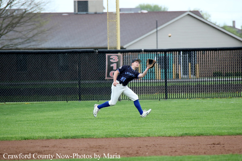 Baseball player in navy jersey and white pants runs in the outfield with his glove up as a ball sails overhead.