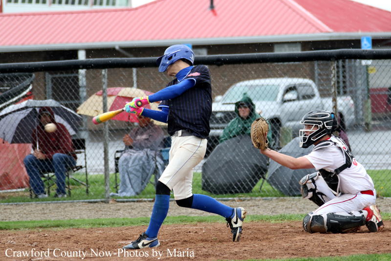 Young baseball batter in blue uniform swings at a pitch while catcher in white gear kneels behind home plate on a dirt field, with spectators in the background.