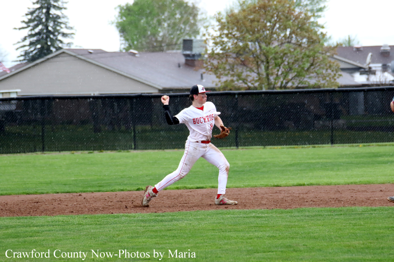 Baseball pitcher in a white and red uniform throwing a pitch on the infield, with a chain-link fence and houses in the background.