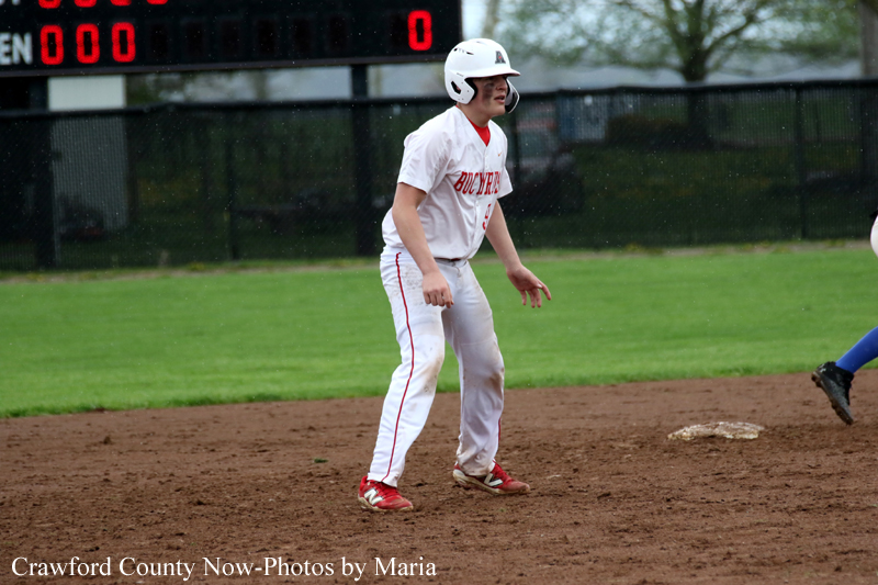 Baseball player in a white uniform and helmet stands on dirt infield during a game, with a fence and scoreboard behind.