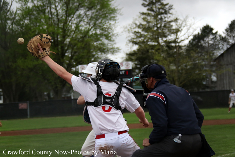 Baseball catcher in white uniform reaches up with his glove as a ball sails by during a play at the plate, with a coach/umpire nearby on the field.