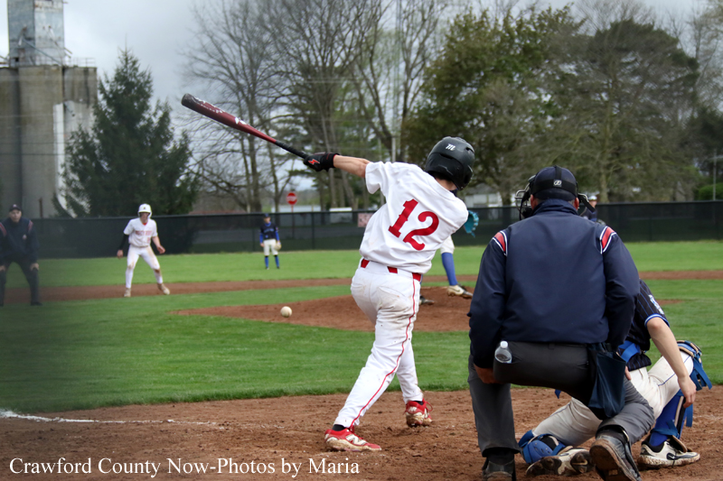 Baseball batter wearing #12 in white with red trim swings at a pitch, with catcher and umpire behind him on the infield.