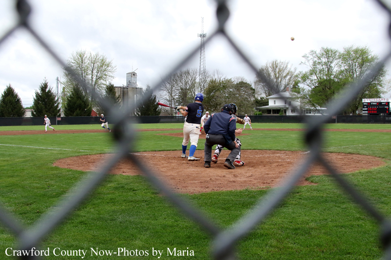 Baseball game seen through a chain-link fence; batter swinging at home plate with catcher and umpire nearby.