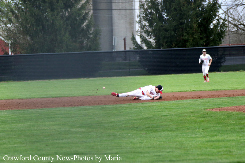 Baseball player slides headfirst on dirt to field a ground ball, with another fielder running in the outfield in the background.
