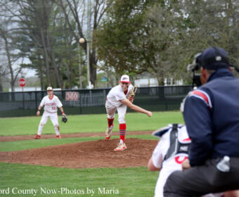 Baseball pitcher in white uniform on the mound delivers a pitch, with a fielder and umpire visible in the foreground on a green field.