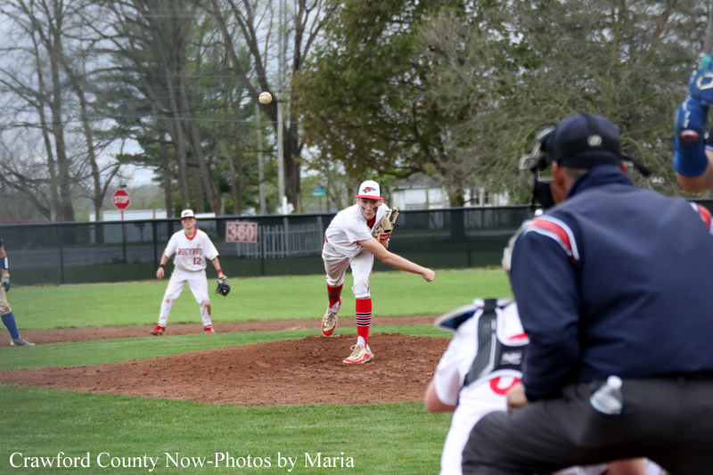 Baseball pitcher in white uniform on the mound delivers a pitch, with a fielder and umpire visible in the foreground on a green field.