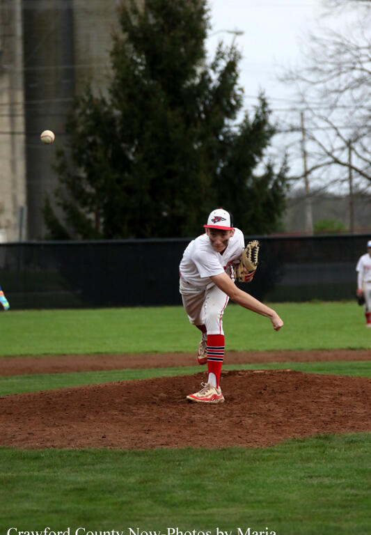 Pitcher in a white uniform on the pitcher's mound delivering a throw during a baseball game when the ball is in the air to the left.