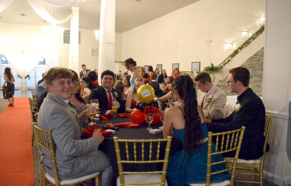 People in formal attire seated around a long banquet table at a decorated event hall, holding drinks and chatting.