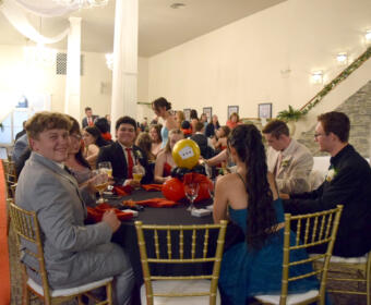 People in formal attire seated around a long banquet table at a decorated event hall, holding drinks and chatting.
