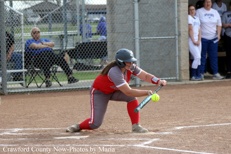 Female softball player in red and gray uniform swings a bat at a pitched ball near home plate, wearing a protective helmet and knee-high red socks as teammates watch nearby.