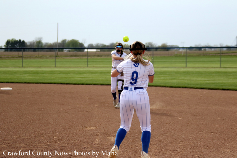 Softball catcher wearing a face mask throws a ball toward a pitcher in a white pinstripe uniform on a dirt infield, with a grassy field and fence in the background.