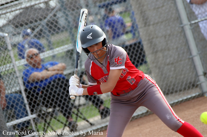 Female softball batter in red and gray swings with a helmet on, bat raised at the plate.