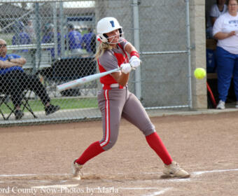 Female softball player in gray-red uniform batting during a game on a dirt field with a chain-link fence in the background.