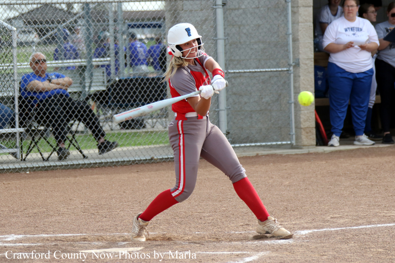 Female softball player in gray-red uniform batting during a game on a dirt field with a chain-link fence in the background.