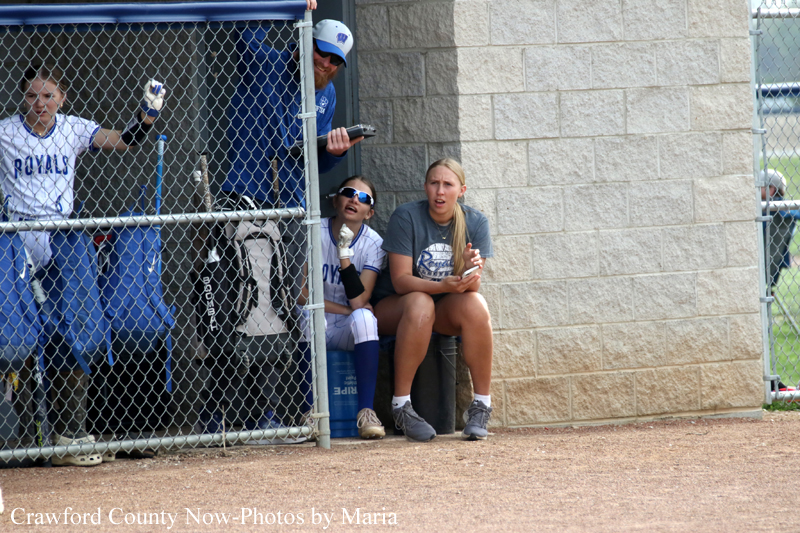 Group of young softball players sitting in a dugout behind a chain-link fence, some in white-blue uniforms, one wearing sunglasses and talking on a phone/light object. The scene is beside a beige brick wall with a man standing in the doorway above them.