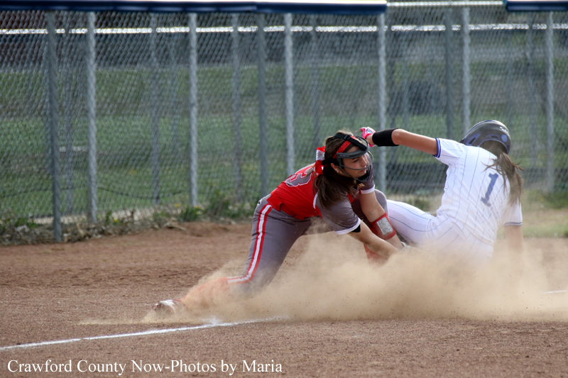 Softball action: a red-uniform runner slides into a base while a white-uniform fielder reaches to tag, dust rising from the dirt.