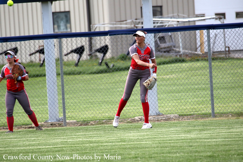 Two female softball players in red and gray uniforms stand near a chain-link outfield fence during a game, one ready to field a ball and the other in a ready position to catch.