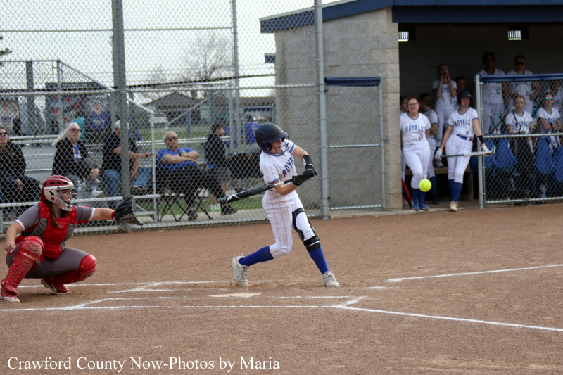 Female softball batter in a white and blue uniform swings at a pitch at home plate, with a red-gear catcher and dugout spectators in the background.