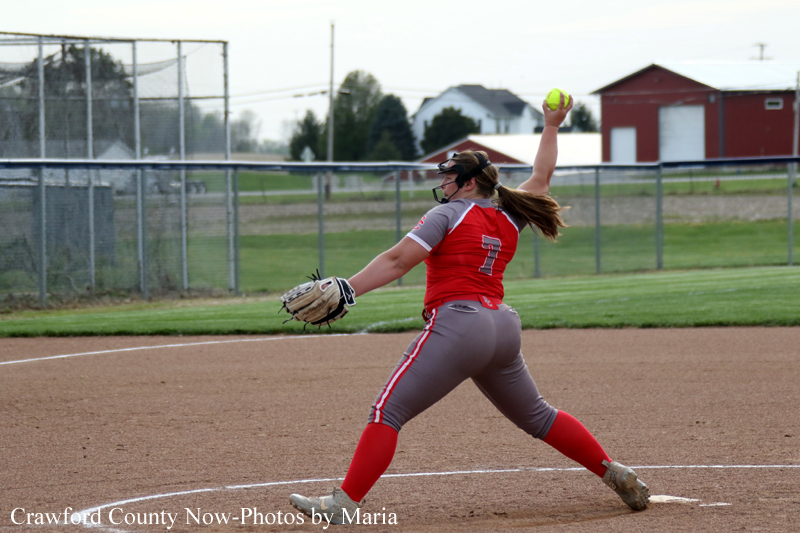 Female softball pitcher in red and gray uniform (number 7) winds up to throw on the dirt mound.