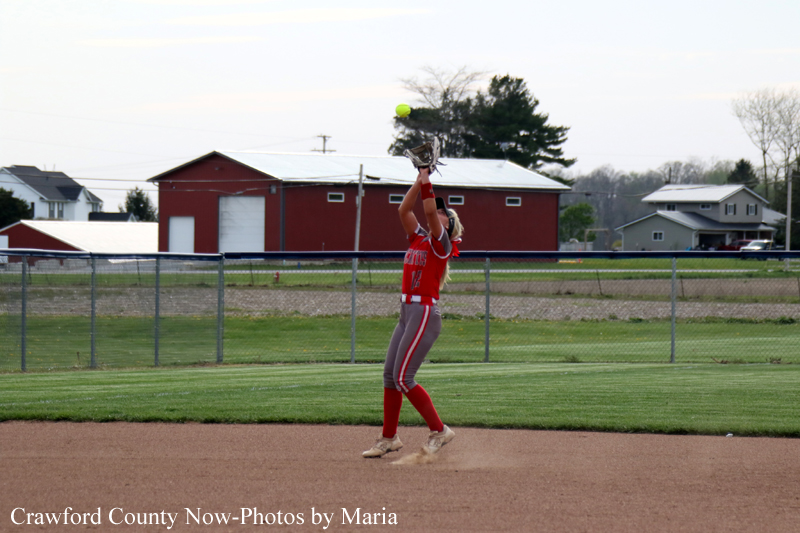 Softball player in a red uniform leaps to catch a ball above her glove on the infield dirt, with a fencing and buildings in the background.