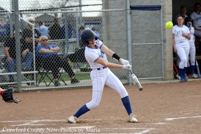Female softball player in white uniform bats at home plate, wearing a helmet and blue socks, mid-swing during a game.