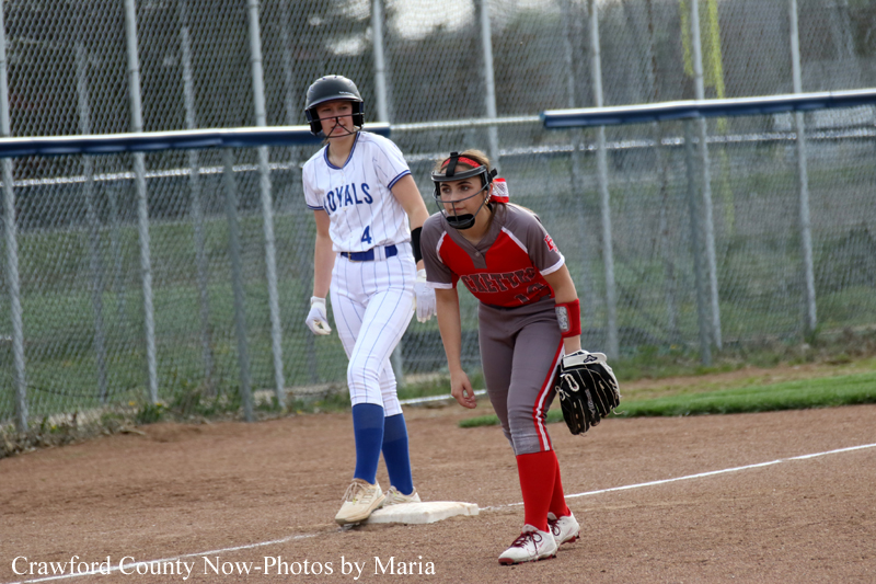 Two female softball players on a dirt infield near a chain-link fence: a baserunner in a white and blue uniform stands on first base while a fielder in red and gray crouches with a glove nearby.