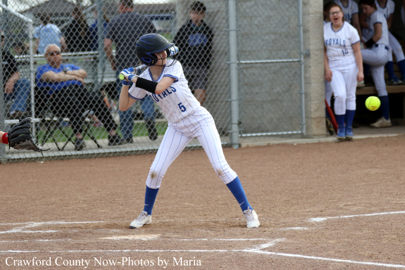 A female softball batter in a white pinstripe uniform and blue helmet swings at a ball on a dirt field, with a chain-link fence and spectators behind her.