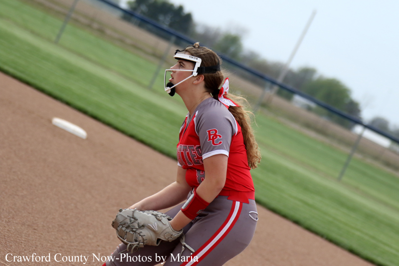 Softball pitcher in a red and gray uniform wearing a protective mask, gripping a glove and preparing to throw on a dirt pitcher's mound.