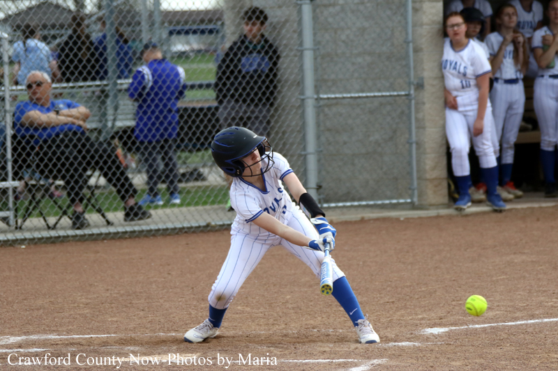 Young softball batter in a white pinstripe uniform swings at a pitch at home plate during a game, helmet on and glove ready.