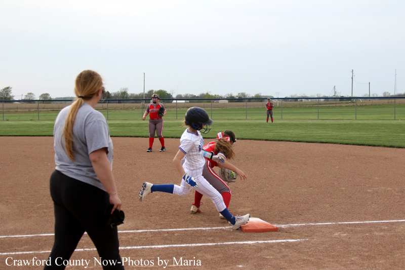 A softball runner slides into first base as a fielder reaches for the ball near the orange base; umpire/coach and another player watch from behind on the infield dirt.