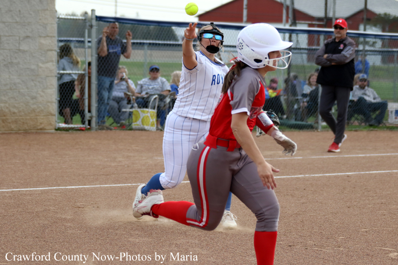 Softball player in white pinstripes slides toward a base while another fielder in gray/red reaches for a ball in the air near the plate; spectators in the background.
