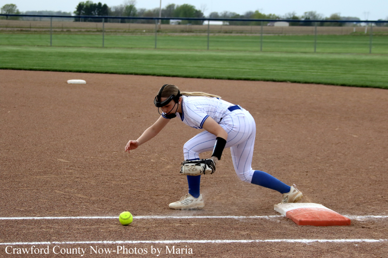 Softball infielder in a white uniform with blue socks bends to field a ground ball near first base.
