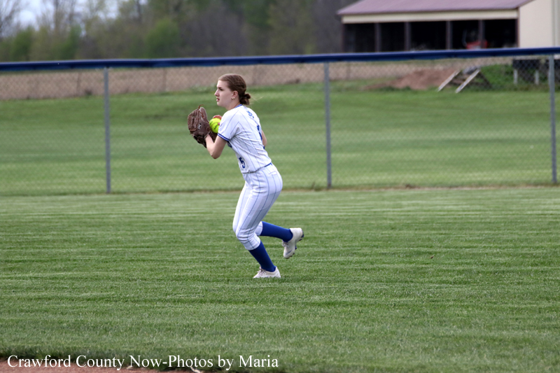 Female softball player in a white uniform with blue socks gloved and ready to catch a ball on a grassy field, with a chain-link fence in the background.