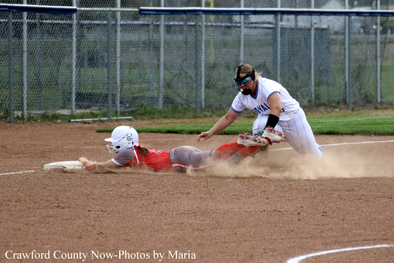 Baseball/softball action: runner in red slides into a base as a fielder in white reaches for the ball, dust rising from the dirt.