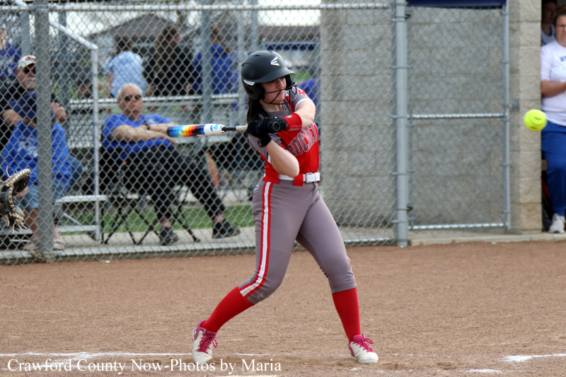 Female softball player in a red and gray uniform swings a bat during a game, wearing a helmet and standing at home plate on a dirt field beside a chain-link fence.