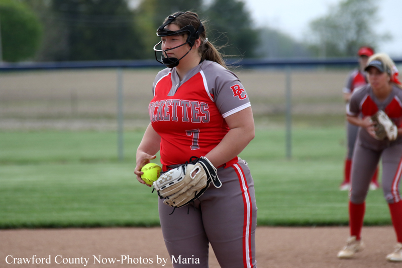 Female softball player in red and gray uniform with number 1, wearing a protective mask, holds a glove and bright yellow ball on a field with teammates in the background.