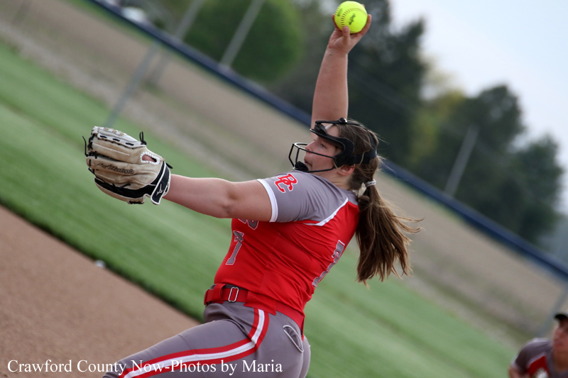 Female softball pitcher in red and gray uniform winds up to throw a pitch on the dirt mound in a game.