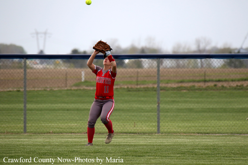 Softball player in red uniform (number 11) catches a ball reaching above the glove near the outfield fence.