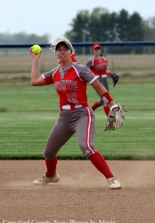 Female softball pitcher in a red-and-gray uniform (number 12) throws a pitch on a dirt infield, with a fielder in the background.","A softball pitcher in red jersey and gray pants winds up a throw on the field while another player watches in the distance.","Action shot of a female softball player mid-pitch on the infield, wearing red and gray with a glove on her left hand.","Pitcher in a red uniform hurls a ball from the mound during a game, with teammates visible in the background.","A female softball player wearing number 12 prepares to throw, standing on the dirt of the pitcher's circle."