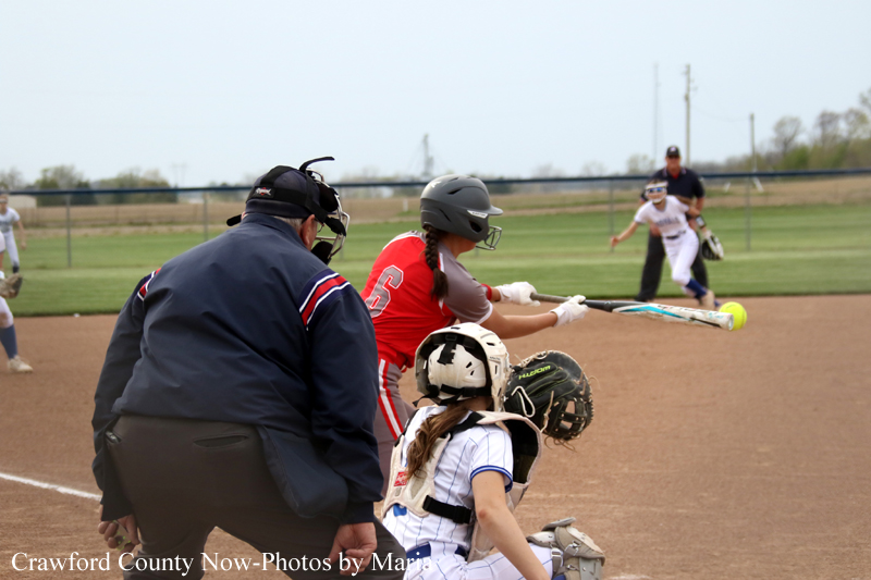 A softball batter in red swings at a pitch as the catcher and umpire stand behind home plate on a dirt field.