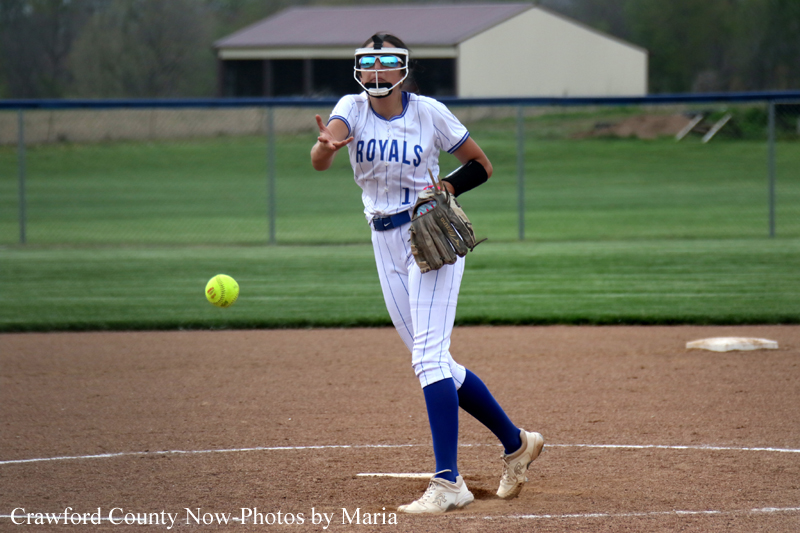 Softball player in a Royals jersey throws a ball on the infield, wearing a helmet and glove.