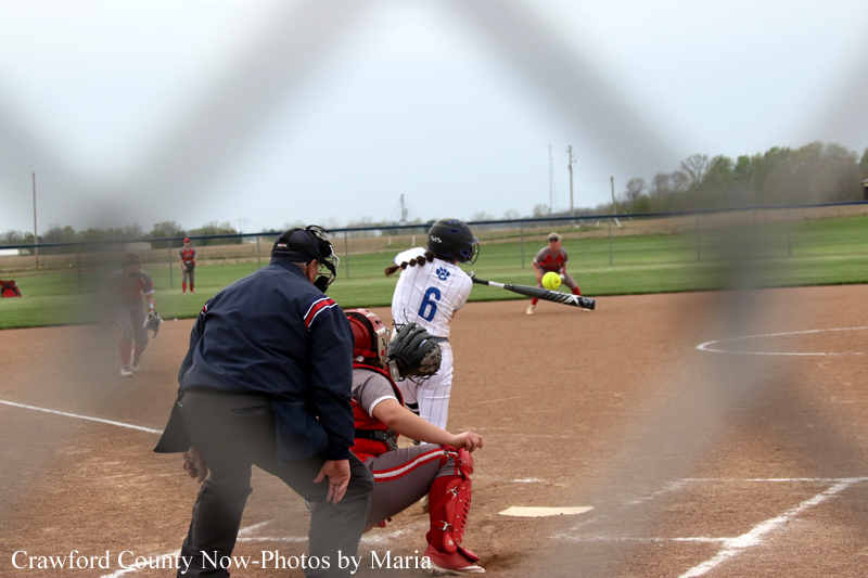 A batter at bat swinging at a softball pitched toward home plate, with catcher and umpire behind, during a game on a dusty infield.