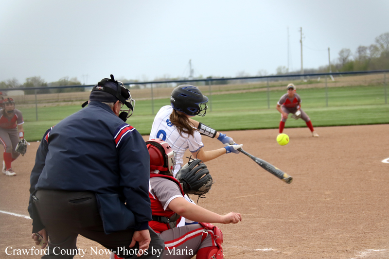 Batter in white and blue swings at a yellow softball as the catcher and umpire watch from home plate on a dirt infield.
