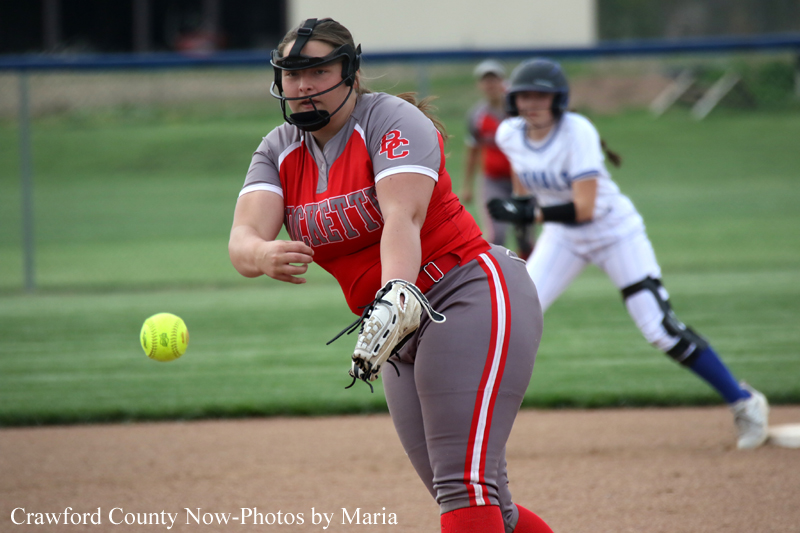 Softball pitcher in red and gray uniform winds up to throw, ball in midair with a fielder in white in the background.