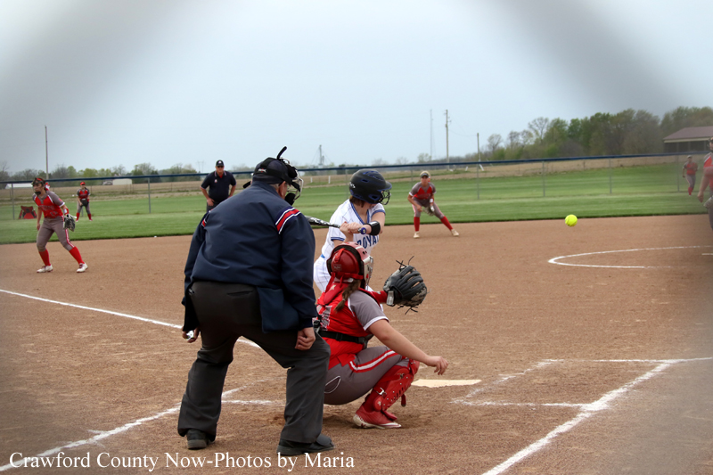Softball batter swings at a pitched ball as the catcher crouches at home plate and the umpire watches nearby.