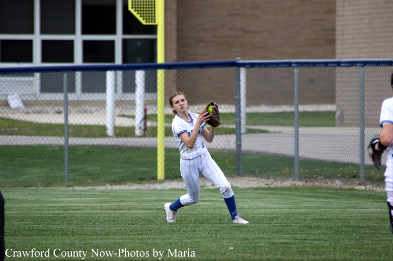 Female softball player in a white-and-blue uniform catches a ground ball with her glove on a grassy field beside a chain-link fence.