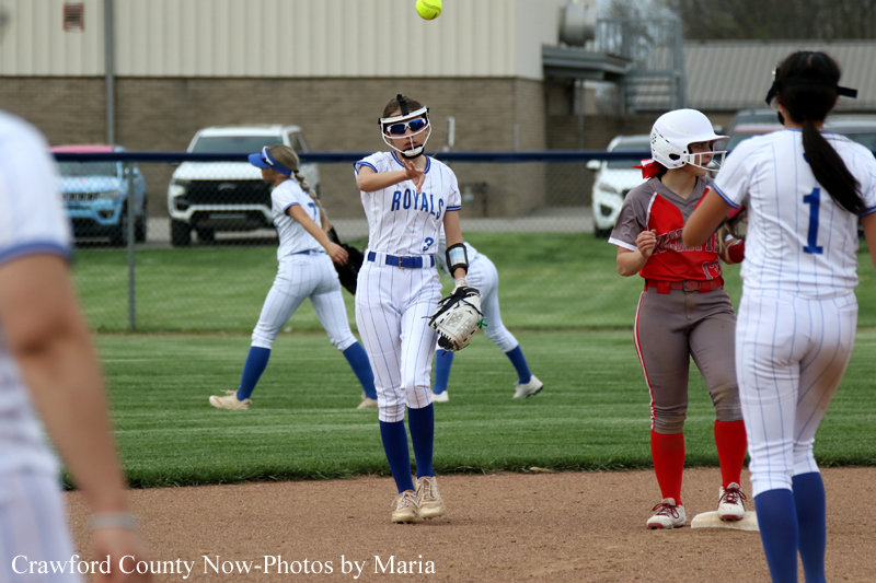 Softball game in progress: pitcher in a Royals uniform throws a pitch, with teammates and opponents nearby on the infield.